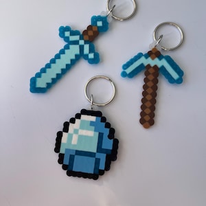 May include: Three pixelated keyrings: a sword, a pickaxe, and a diamond. The sword is light blue with a brown handle. The pickaxe is brown and light blue. The diamond is light and dark blue with a black outline. All have silver key rings.