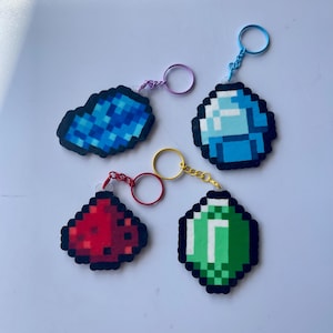 Könnte beinhalten: Vier pixelige Schlüsselanhänger im Bild. Einer ist blau, einer rot, einer grün und einer blau und weiß. Jeder Schlüsselanhänger hat einen unterschiedlich farbigen Ring und eine Kette. Die Schlüsselanhänger sind wahrscheinlich aus Kunststoffperlen gefertigt.