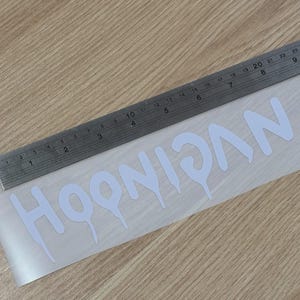 May include: White "HOONIGAN" decal with a dripping font style, placed on a clear backing. A metal ruler is positioned above the decal, showing inch measurements. The decal is on a light wood surface.