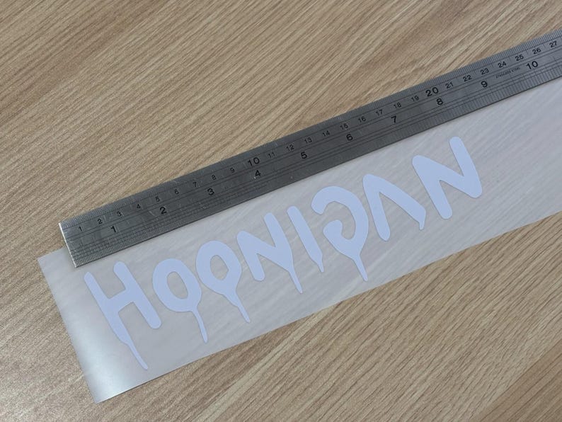 May include: White "HOONIGAN" decal with a dripping font style, placed on a clear backing. A metal ruler is positioned above the decal, showing inch measurements. The decal is on a light wood surface.