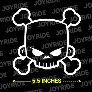 May include: A white vinyl decal featuring a stylized skull and crossbones design with sharp, angular eyes and a toothy grin. The decal is positioned above a yellow measurement line labeled 5.5 INCHES, indicating its width.
