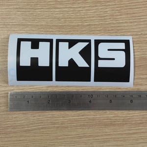 May include: A rectangular white sticker with the letters "HKS" in white, set against a black background. The sticker is placed on a light brown surface next to a metal ruler. The ruler measures approximately 6 inches.