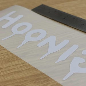 May include: White vinyl decal with the word "HOONIGANS" in a bold, stylized font. A metal ruler is positioned near the decal on a light wood surface. The decal is on a transparent backing.