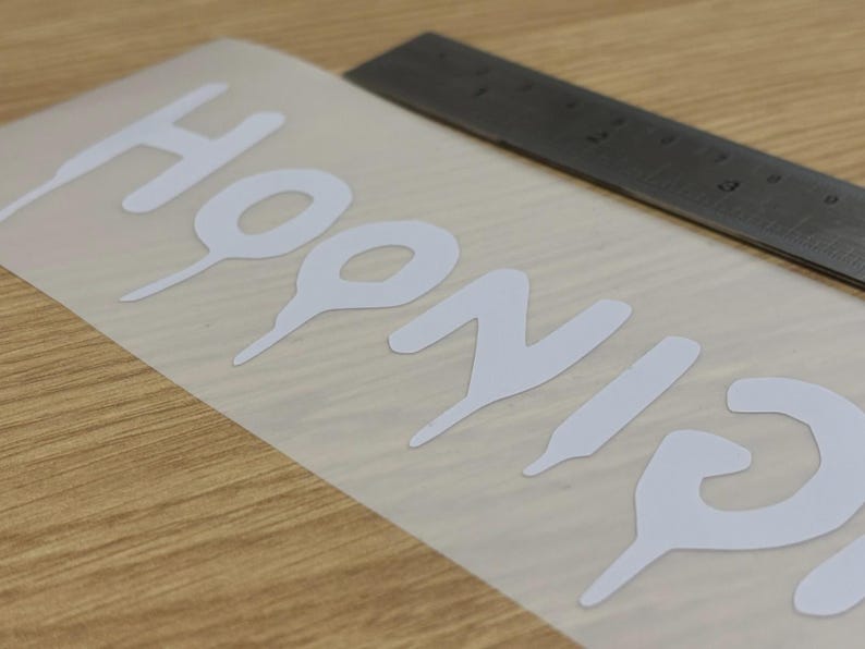 May include: White vinyl decal with the word "HOONIGANS" in a bold, stylized font. A metal ruler is positioned near the decal on a light wood surface. The decal is on a transparent backing.