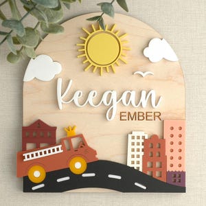 May include: Wooden name plaque with a city theme. Features a yellow sun, white clouds, and a fire truck. The name "Keegan Ember" is displayed in white script. The background is a light wood color.