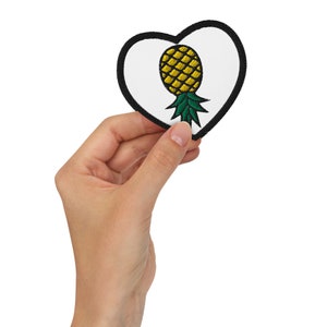 Puede incluir: Un parche blanco en forma de corazón con un contorno negro y una piña amarilla bordada con hojas verdes.