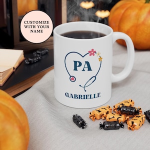 Könnte beinhalten: Eine weiße Kaffeetasse mit einem blauen Stethoskop und einem Herzdesign. Der Text "PA" befindet sich im Herzen. Der Text "Gabrielle" befindet sich unter dem Herzen. Die Tasse steht auf einer weißen Oberfläche mit orangefarbenen und schwarzen Süßigkeiten.