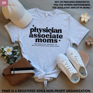 May include: A light gray t-shirt with the text "physician associate moms" in black. The shirt is styled with a white baseball cap, white sneakers, a brown purse, and decorative flowers. The text "Empowering Women in Motherhood and Medicine" is also on the shirt.
