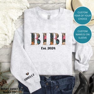 May include: Gray sweatshirt with the text "BIBI Est. 2024" in black with floral accents. The left sleeve has the name "GISELLE" in black with a heart.