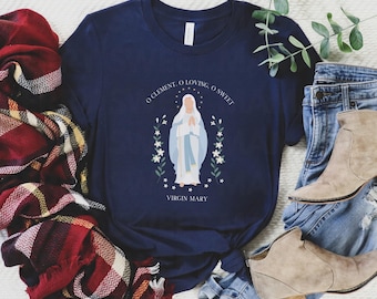 以下が含まれることがあります： 聖母マリアのグラフィックが特徴のネイビーブルーのTシャツ。「O Clement, O Loving, O Sweet」と「Virgin Mary」の文字が白でデザインされています。グラフィックは白い花と緑の葉で囲まれています。Tシャツはブルージーンズとタンブーツと組み合わせています。