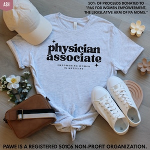 May include: A light gray t-shirt with the words "physician associate" in black, with the text "Empowering Women in Medicine" below. The shirt is paired with white sneakers, a white cap, and a brown purse. The text on the shirt is the focal point.