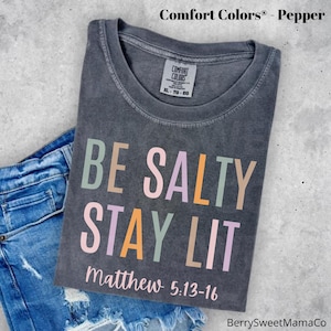 May include: A gray t-shirt with the text "BE SALTY STAY LIT" in colorful letters. The text "Matthew 5:13-16" is printed below the main text.