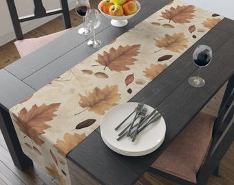 May include: A beige table runner with a repeating pattern of brown autumn leaves and acorns. The runner is placed on a dark gray wooden table set for a meal, with plates, forks, and wine glasses. A bowl of apples sits in the center.