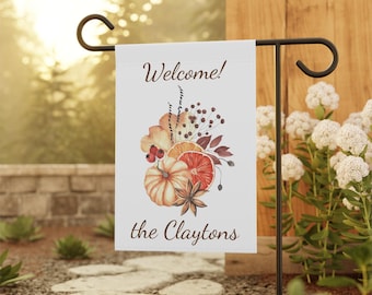 Może przedstawiać: Biała flaga ogrodowa z napisem "Welcome!" i "the Claytons" w brązowym skrypcie. Flaga przedstawia akwarelową ilustrację dyni, plasterków pomarańczy i jesiennych liści. Flaga jest wyświetlana na czarnym metalowym paliku ogrodowym.
