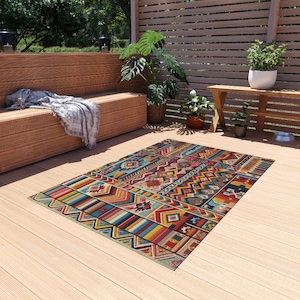 Puede incluir: Una alfombra rectangular para exteriores con un vibrante patrón geométrico. La alfombra presenta una mezcla de colores como rojo, azul, amarillo y turquesa, con varias formas y diseños. La alfombra está colocada sobre una terraza de madera.