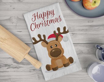 May include: White Christmas kitchen towel with a cartoon reindeer wearing a Santa hat. The text "Happy Christmas" is written in red script. A wooden rolling pin and two apples on a plate are in the background.