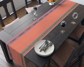 Op de afbeelding: Een koraalkleurige tafelloper met een gestreept patroon en decoratieve randen in blauw, groen en bruin. De loper ligt op een donkere houten tafel die gedekt is voor een maaltijd, met borden, bestek en wijnglazen.