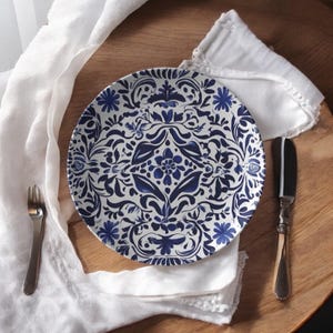 May include: A white and blue decorative plate with a floral pattern. The plate is set with a white napkin, a fork, and a knife on a wooden surface. The plate's design includes blue flowers and leaves.