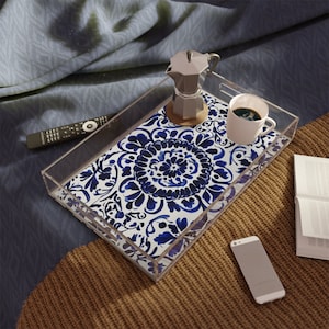 May include: A clear acrylic tray with a blue and white floral design. The tray contains a silver coffee maker, a white coffee cup, a remote control, a smartphone, and an open book. The tray rests on a brown knitted surface.