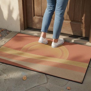 Könnte beinhalten: Eine rechteckige Fußmatte mit einem Sonnenuntergangsdesign in Pfirsich-, Gelb- und Beigetönen. Die Matte hat einen schwarzen Rand und liegt vor einer Holztür. Das Design zeigt eine große Sonne und horizontale Streifen.
