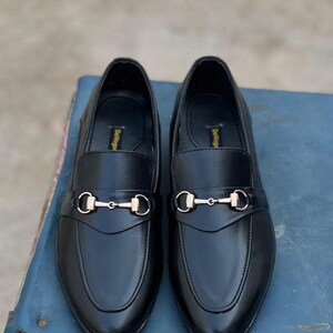 Pu&ograve; includere: Un paio di mocassini in pelle nera con fibbia in metallo color oro. Le scarpe sono esposte su una superficie blu. Le scarpe hanno un design classico, adatto per abbigliamento formale o casual.