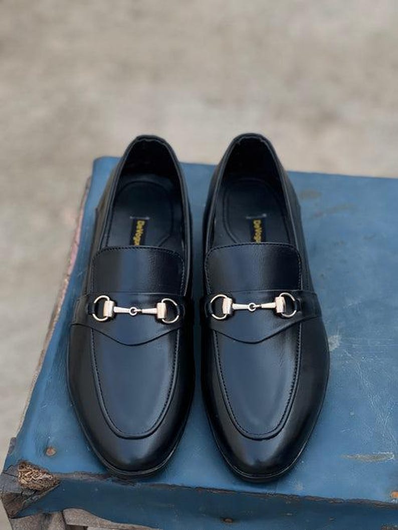 Pu&ograve; includere: Un paio di mocassini in pelle nera con fibbia in metallo color oro. Le scarpe sono esposte su una superficie blu. Le scarpe hanno un design classico, adatto per abbigliamento formale o casual.