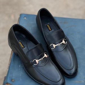 Pu&ograve; includere: Un paio di mocassini in pelle nera con dettagli dorati a forma di morsetto. Le scarpe hanno un design classico con punta arrotondata e tacco basso. Il marchio "DeVogue" &egrave; visibile sulla soletta. Su una superficie blu.