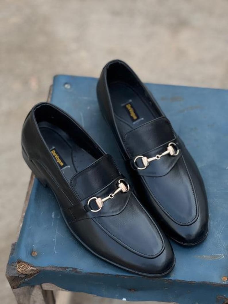 Pu&ograve; includere: Un paio di mocassini in pelle nera con dettagli dorati a forma di morsetto. Le scarpe hanno un design classico con punta arrotondata e tacco basso. Il marchio "DeVogue" &egrave; visibile sulla soletta. Su una superficie blu.