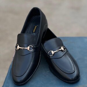 Pu&ograve; includere: Un paio di mocassini in pelle nera con finiture in metallo dorato. Le scarpe hanno un design classico con punta arrotondata e tacco basso. I mocassini sono esposti su una superficie blu.