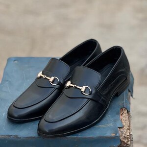 Pu&ograve; includere: Un paio di mocassini in pelle nera con dettagli metallici color oro. Le scarpe presentano un design classico con punta arrotondata e cinturino decorativo. I mocassini sono esposti su una superficie blu.