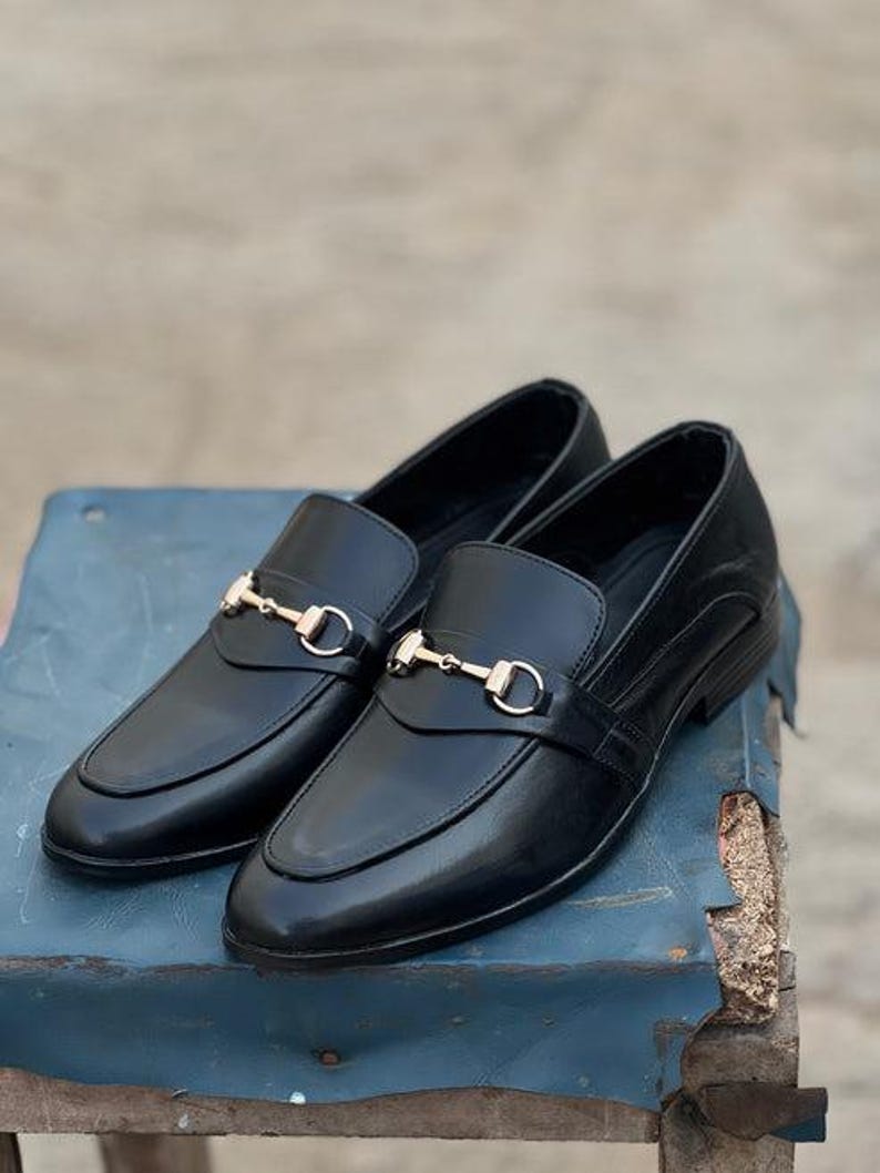 Pu&ograve; includere: Un paio di mocassini in pelle nera con dettagli metallici color oro. Le scarpe presentano un design classico con punta arrotondata e cinturino decorativo. I mocassini sono esposti su una superficie blu.