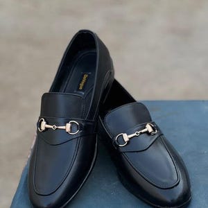 Pu&ograve; includere: Un paio di mocassini in pelle nera con dettagli metallici color oro. Le scarpe hanno un design classico con punta arrotondata e cinturino sulla tomaia. I mocassini sono esposti su una superficie blu.