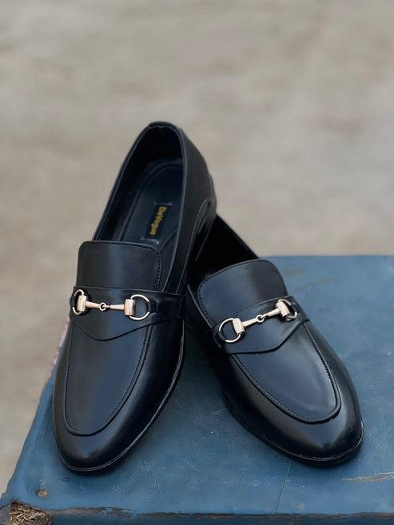 Pu&ograve; includere: Un paio di mocassini in pelle nera con dettagli metallici color oro. Le scarpe hanno un design classico con punta arrotondata e cinturino sulla tomaia. I mocassini sono esposti su una superficie blu.