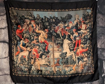 May include: A tapestry depicting a hunting scene with a unicorn, hunters, and dogs in a forest setting. The tapestry is framed with a black border and features a gold trim.