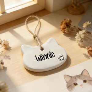 May include: A white cat-shaped ornament with the name "Winnie" in black script and a small star graphic. The ornament hangs from a natural twine loop. The background includes dried flowers and a cat illustration.