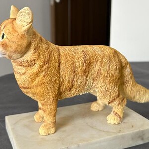 Puede incluir: Una figurita de cer&aacute;mica de un gato jengibre de pie sobre una base de m&aacute;rmol blanco. El gato est&aacute; representado en un estilo realista con pelo y rasgos faciales detallados.