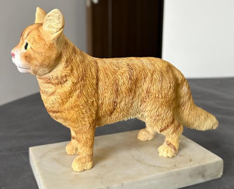 Puede incluir: Una figurita de cer&aacute;mica de un gato jengibre de pie sobre una base de m&aacute;rmol blanco. El gato est&aacute; representado en un estilo realista con pelo y rasgos faciales detallados.