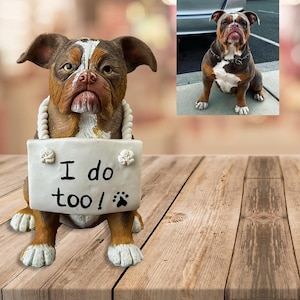 May include: A brown and white bulldog figurine holding a sign that says "I do too!" with a paw print. The figurine is sitting on a wooden surface.
