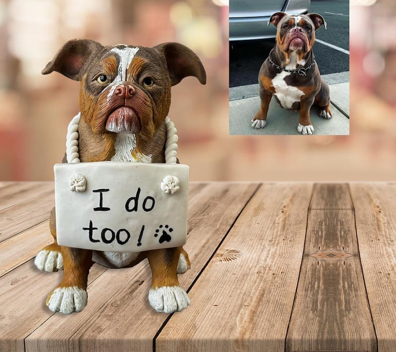 May include: A brown and white bulldog figurine holding a sign that says "I do too!" with a paw print. The figurine is sitting on a wooden surface.