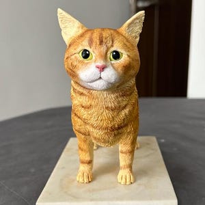 Puede incluir: Una figurita realista de un gato jengibre de pie sobre una base de m&aacute;rmol blanco. El gato tiene ojos verdes y una mancha blanca en el pecho.