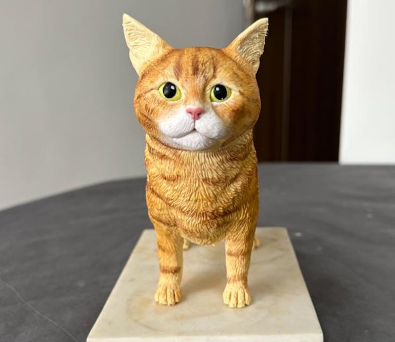 Puede incluir: Una figurita realista de un gato jengibre de pie sobre una base de m&aacute;rmol blanco. El gato tiene ojos verdes y una mancha blanca en el pecho.