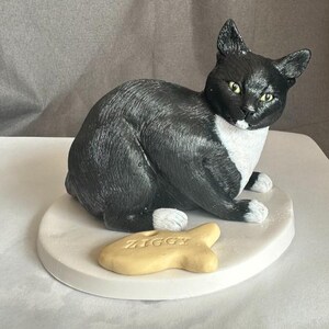 Puede incluir: Una figurita de gato negro y blanco con una mancha blanca en el pecho, sentada en un pedestal blanco con una golosina amarilla en forma de pez que dice "Ziggy".