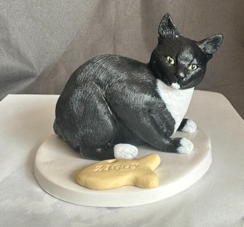 Puede incluir: Una figurita de gato negro y blanco con una mancha blanca en el pecho, sentada en un pedestal blanco con una golosina amarilla en forma de pez que dice "Ziggy".
