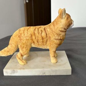 Puede incluir: Una figurita de un gato jengibre de pie sobre una base de m&aacute;rmol blanco. El gato est&aacute; representado en un estilo realista con pelo detallado y una postura natural.
