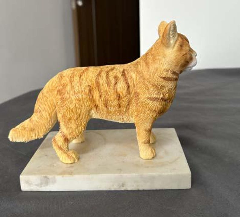 Puede incluir: Una figurita de un gato jengibre de pie sobre una base de m&aacute;rmol blanco. El gato est&aacute; representado en un estilo realista con pelo detallado y una postura natural.