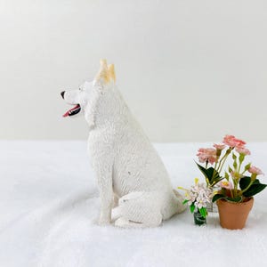 May include: White ceramic figurine of a sitting German Shepherd dog with a light brown patch on its head. The dog is on a white surface with a small potted plant with pink flowers in the background.