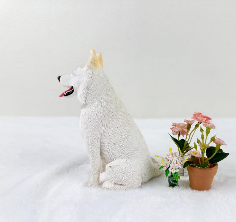 May include: White ceramic figurine of a sitting German Shepherd dog with a light brown patch on its head. The dog is on a white surface with a small potted plant with pink flowers in the background.