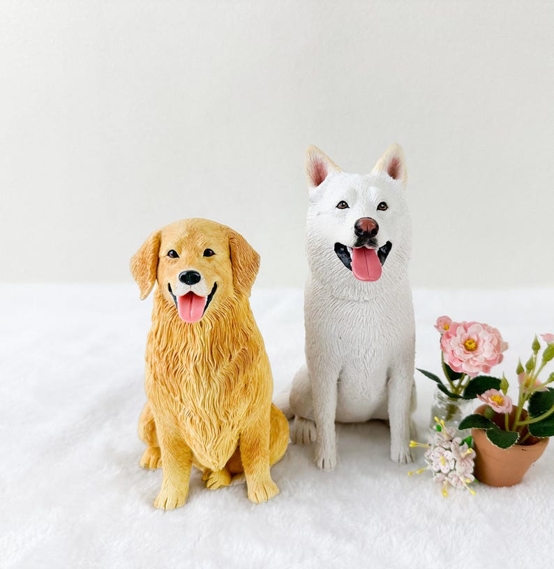Puede incluir: Dos figuritas de perros realistas, un golden retriever y un perro blanco, ambos sentados con la lengua fuera. El golden retriever tiene la nariz marr&oacute;n y el perro blanco tiene la nariz negra. Las figuritas est&aacute;n sobre una superficie blanca con una peque&ntilde;a planta en maceta en el fondo.