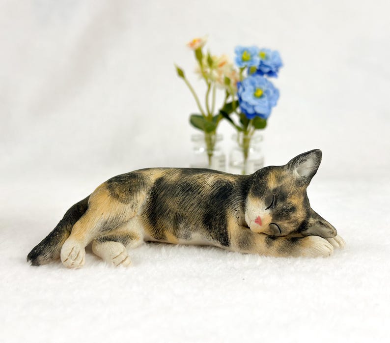 Puede incluir: Una figurita de gato calico durmiendo sobre una superficie blanca. El gato est&aacute; acostado de lado con la cabeza apoyada en sus patas. El gato tiene pelo negro, marr&oacute;n y blanco.