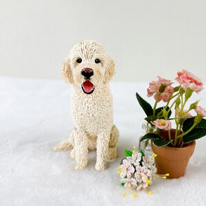 May include: A white cream-colored dog figurine with a brown nose and a pink tongue sticking out. The dog is sitting with its paws crossed and is looking directly at the camera. The figurine is on a white surface with a small bouquet of pink and white flowers in the foreground.